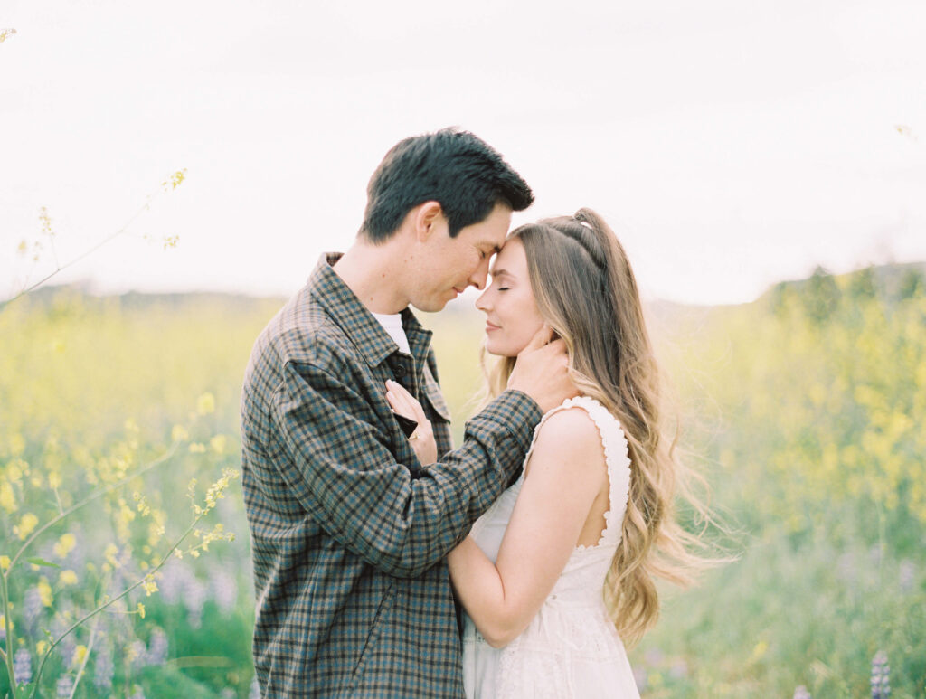 Malibu Engagement Session - Malibu Canyon - A couple stands closely in a field of wildflowers. The man gently holds the woman’s face as they touch foreheads, eyes closed, sharing a tender moment. The background is soft and serene with greenery and yellow blooms. Copyright - Lisa Leanne Photography, California