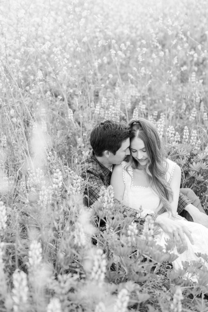 Malibu Engagement Session - Malibu Canyon - A couple sits closely together in a field of wildflowers, smiling and sharing an intimate moment. The photo is in black and white, creating a soft, romantic atmosphere. Copyright - Lisa Leanne Photography, California