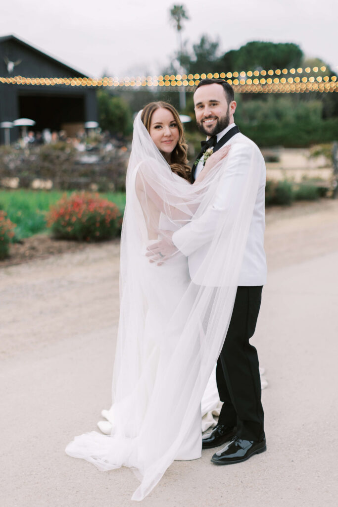A bride and groom stand close together outdoors, smiling at the camera. The bride wears a white dress with a long veil, and the groom wears a white tuxedo jacket with black pants. String lights and greenery are in the background. Copyright - Lisa Leanne Photography, California