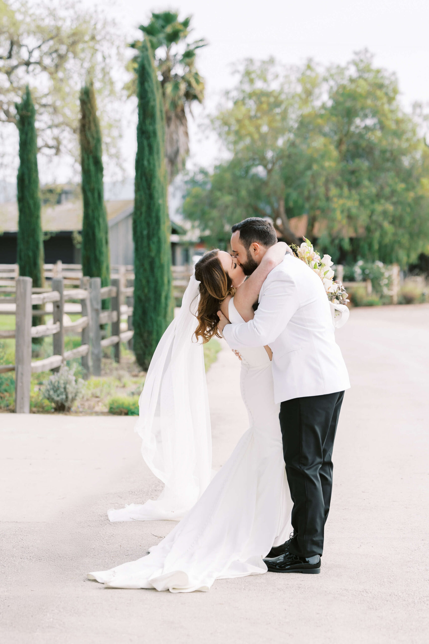 A bride and groom embrace and kiss outdoors on a sunny day. The bride wears a white dress and veil, holding a bouquet, while the groom wears a white jacket and black pants. Trees and a wooden fence line the background. Copyright - Lisa Leanne Photography, California