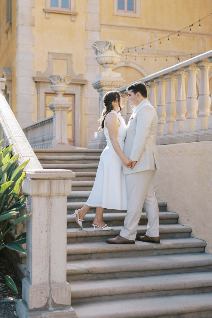 Villa Del Sol D'Oro - A couple in light-colored formal attire stands on outdoor stone steps, holding hands and facing each other, with a historic building and plants in the background. The scene is bright and romantic. Copyright - Lisa Leanne Photography, California