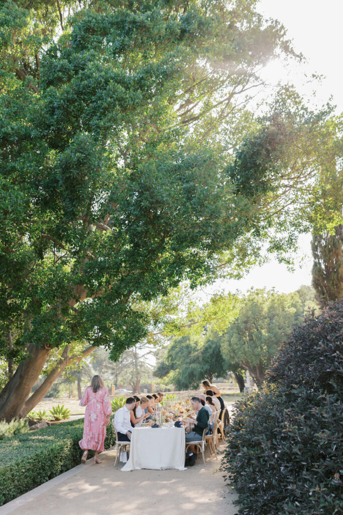 Villa Del Sol D'Oro Rehearsal Dinner - A group of people sit around a table enjoying an outdoor meal under a large, leafy tree on a sunny day, while a woman in a pink dress approaches the table along a garden path. Copyright - Lisa Leanne Photography, California