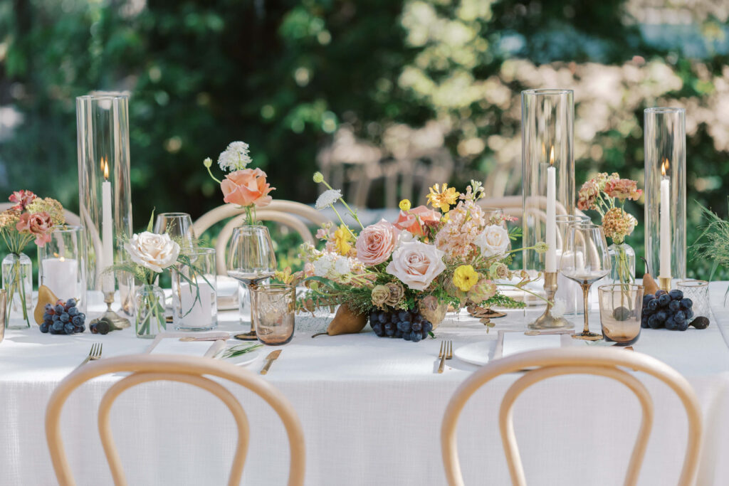 Villa Del Sol D'Oro - Elegant outdoor table setting with a white tablecloth, floral centerpieces, tall glass candle holders, glassware, small bunches of grapes, and light wooden chairs, surrounded by greenery in the background. Copyright - Lisa Leanne Photography, California